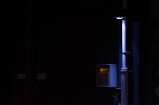 A dimly lit scene featuring a vertical pipe and an electrical panel with glowing indicator lights. The surrounding environment is mostly dark, highlighting the contrast with the illuminated objects.