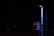 A dimly lit scene featuring a vertical pipe and an electrical panel with glowing indicator lights. The surrounding environment is mostly dark, highlighting the contrast with the illuminated objects.