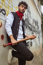 A young man with curly hair stands against a graffiti-covered wall, holding a large wooden bat. He wears a black varsity jacket over a white shirt, a red bandana around his neck, and white sneakers. The atmosphere around him is urban, with a casual yet confident demeanor.