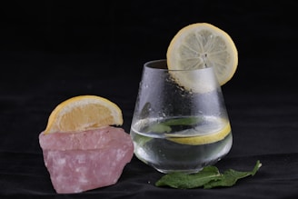A glass of water with a lemon slice on its rim sits beside a pink rock with another lemon slice on top. There are also a few mint leaves scattered nearby on a dark surface, creating an elegant and refreshing aesthetic.