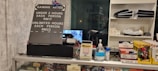 The image displays a counter with various items including a pricing sign for gaming rates, a payment terminal, hand sanitizer, a scanner, and shelves with playing cards and Lego sets. There is a screen showing what appears to be a security camera feed.