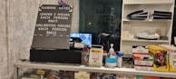 The image displays a counter with various items including a pricing sign for gaming rates, a payment terminal, hand sanitizer, a scanner, and shelves with playing cards and Lego sets. There is a screen showing what appears to be a security camera feed.