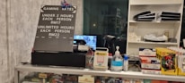 The image displays a counter with various items including a pricing sign for gaming rates, a payment terminal, hand sanitizer, a scanner, and shelves with playing cards and Lego sets. There is a screen showing what appears to be a security camera feed.