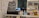 The image displays a counter with various items including a pricing sign for gaming rates, a payment terminal, hand sanitizer, a scanner, and shelves with playing cards and Lego sets. There is a screen showing what appears to be a security camera feed.