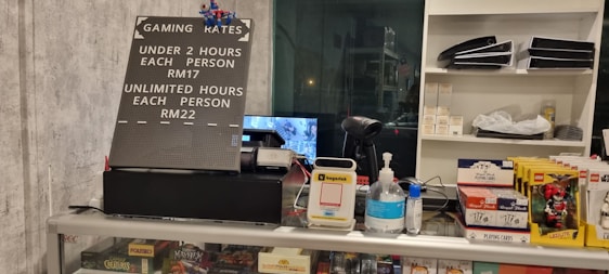 The image displays a counter with various items including a pricing sign for gaming rates, a payment terminal, hand sanitizer, a scanner, and shelves with playing cards and Lego sets. There is a screen showing what appears to be a security camera feed.