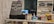 The image displays a counter with various items including a pricing sign for gaming rates, a payment terminal, hand sanitizer, a scanner, and shelves with playing cards and Lego sets. There is a screen showing what appears to be a security camera feed.