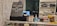 The image displays a counter with various items including a pricing sign for gaming rates, a payment terminal, hand sanitizer, a scanner, and shelves with playing cards and Lego sets. There is a screen showing what appears to be a security camera feed.