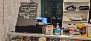 The image displays a counter with various items including a pricing sign for gaming rates, a payment terminal, hand sanitizer, a scanner, and shelves with playing cards and Lego sets. There is a screen showing what appears to be a security camera feed.