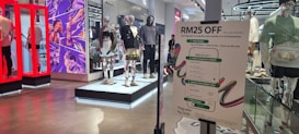 The image depicts the interior of a clothing store with multiple mannequins dressed in various outfits. There is a prominent sign advertising a discount of RM25 off with specific payment conditions. The store features modern and futuristic design elements, including vibrant red display frames and a comic-style mural on the wall.