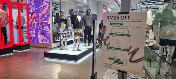 The image depicts the interior of a clothing store with multiple mannequins dressed in various outfits. There is a prominent sign advertising a discount of RM25 off with specific payment conditions. The store features modern and futuristic design elements, including vibrant red display frames and a comic-style mural on the wall.