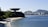 An iconic, futuristic building resembling a flying saucer is perched on a cliff overlooking a sandy beach. Lush greenery surrounds the area, and in the background, a cityscape with tall buildings is visible against mountainous terrain. The sea in the foreground reflects a calm, clear blue under a bright sky.