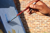 A hand holding a paintbrush is applying blue paint to a canvas that has a sketched design. The background consists of a brick wall, and sunlight casts shadows on the canvas and hand.