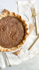 A freshly baked pie with a swirled top sits on a white ceramic dish. It's placed on a wooden board surrounded by torn book pages. A gold fork rests beside the pie, creating a simple and elegant setting.