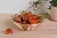 A wicker basket full of large, dried, orange, and brown fungi sits on a light wooden surface. A single dried mushroom lies on the table next to the basket, and a vine plant in a white pot is visible in the background.