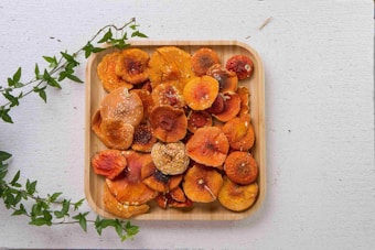 A wooden tray filled with dried, orange mushrooms placed on a textured white surface. Fresh green vine leaves partially frame the tray, adding a touch of natural color contrast.