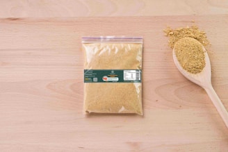 A sealed transparent plastic bag filled with brownish powder lies on a wooden surface. Next to it, a wooden spoon holds a small heap of the same powder, with some scattered around. The bag has a label with text and a barcode.
