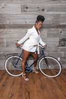 A person dressed in a stylish white blazer and shorts stands beside a blue bicycle against a rustic wooden background. The individual is wearing high-heeled sandals and appears poised and confident.
