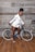A person dressed in a stylish white blazer and shorts stands beside a blue bicycle against a rustic wooden background. The individual is wearing high-heeled sandals and appears poised and confident.