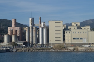 An industrial facility with tall silos and large rectangular buildings near a body of water. The area is surrounded by hills and mountains under a clear blue sky.