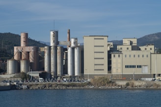 An industrial facility with tall silos and large rectangular buildings near a body of water. The area is surrounded by hills and mountains under a clear blue sky.