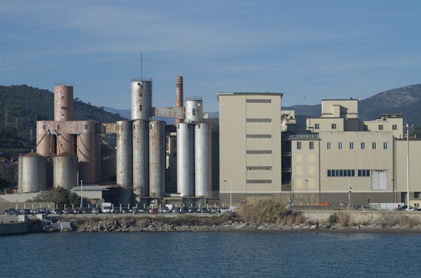 An industrial facility with tall silos and large rectangular buildings near a body of water. The area is surrounded by hills and mountains under a clear blue sky.