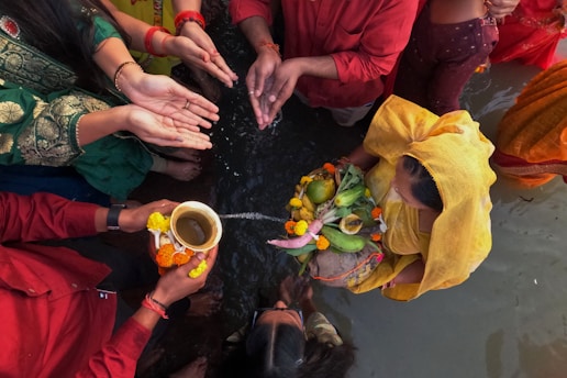Several people are participating in a traditional ritual near water, with hands extended for offerings. They are dressed in colorful garments, and one person holds a decorated plate with fruits and flowers.