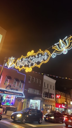A vibrant street scene featuring a colorful array of lights forming the words 'India Square', suspended above the street. Below, a row of shops and restaurants line the street, with visible signs including 'Curry On' and a lounge bar. Cars are parked along the street, and the area is lively and well-lit, suggesting a bustling urban environment.