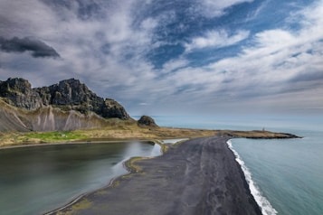 A dramatic landscape featuring a rugged mountain range with craggy peaks under a partially cloudy sky. In the foreground, there is a black sand beach and a calm body of water adjacent to it. To the right, the ocean spreads out toward the horizon with gentle waves. A small red lighthouse is visible on a rocky point, adding a touch of color to the muted tones of the scene.