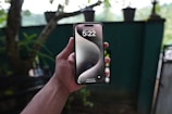 A hand holds a smartphone displaying a lock screen with the time 5:22. The background is a blurred view of a garden with trees and potted plants.