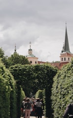 A pathway is surrounded by lush green hedges forming an archway. In the background, architectural spires and ornate towers rise against a cloudy sky. A group of people is gathered along the path, engaging with the surroundings.