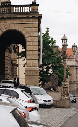 A stone building with an archway is visible next to a row of parked cars. A stone lion statue is perched beside a vintage-style lamppost. Trees and an old building with red roof tiles are in the background, and a person wearing a hat and dark coat is walking towards the cars.