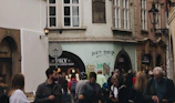 People walking in a narrow street lined with historical buildings, featuring a mix of individuals, some using their phones. Graffiti with the phrase 'JUST DID IT.' is visible on one of the walls. The street includes a shop named VILY advertising exclusive garnet items.