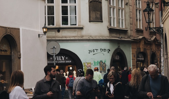 People walking in a narrow street lined with historical buildings, featuring a mix of individuals, some using their phones. Graffiti with the phrase 'JUST DID IT.' is visible on one of the walls. The street includes a shop named VILY advertising exclusive garnet items.