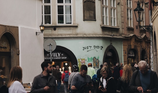 People walking in a narrow street lined with historical buildings, featuring a mix of individuals, some using their phones. Graffiti with the phrase 'JUST DID IT.' is visible on one of the walls. The street includes a shop named VILY advertising exclusive garnet items.