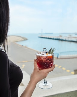 A person is holding a red cocktail adorned with pineapple leaves and an orange slice, while gazing at a calm waterfront. The background features a walkway with barriers and a distant view of boats docked near a pier under a clear sky.