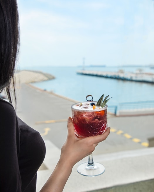A person is holding a red cocktail adorned with pineapple leaves and an orange slice, while gazing at a calm waterfront. The background features a walkway with barriers and a distant view of boats docked near a pier under a clear sky.