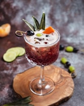 A decorative cocktail in a textured glass sits on a polished wooden coaster. The drink is garnished with a slice of orange peel, a black olive, a green olive, and pineapple leaves. Ice cubes are visible in the dark, frothy liquid. In the background, slices of lime, a tangerine, and a skewer with grapes create a rustic setting.