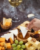 A hand dips a triangular piece of white cheese into a bowl of honey surrounded by various types of cheese, nuts, dried fruits, and grapes. A glass of white wine is visible in the background.
