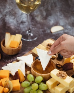 A hand dips a triangular piece of white cheese into a bowl of honey surrounded by various types of cheese, nuts, dried fruits, and grapes. A glass of white wine is visible in the background.