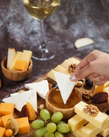 A hand dips a triangular piece of white cheese into a bowl of honey surrounded by various types of cheese, nuts, dried fruits, and grapes. A glass of white wine is visible in the background.