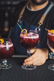 A bartender holding a large glass of a red beverage garnished with an orange slice, a cherry, and a black straw. The person is wearing a dark apron with leather straps and a black shirt. There are two additional similar drinks on either side.