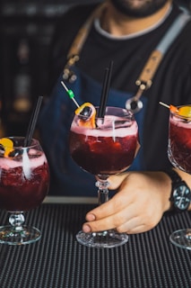 A bartender holding a large glass of a red beverage garnished with an orange slice, a cherry, and a black straw. The person is wearing a dark apron with leather straps and a black shirt. There are two additional similar drinks on either side.
