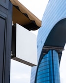 A modern architectural scene featuring a large, sleek skyscraper with reflective blue glass panels. In the foreground, there's a blank white rectangular sign hanging under a brown awning attached to a dark building structure.