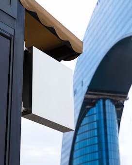 A modern architectural scene featuring a large, sleek skyscraper with reflective blue glass panels. In the foreground, there's a blank white rectangular sign hanging under a brown awning attached to a dark building structure.
