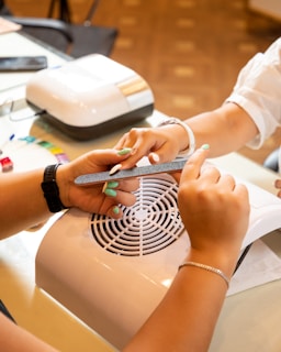 A nail salon setting featuring two hands, one receiving a manicure while the other is filing the nails. The workspace includes a ventilation device and a selection of colored nail polish samples.