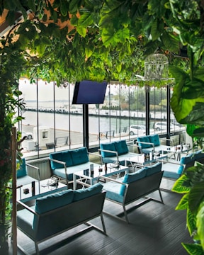 A modern outdoor café with vibrant green foliage hanging from the ceiling, offering a lush atmosphere. The area is furnished with sleek, blue-cushioned seating and glossy tables. Large glass windows provide a view of the waterfront and vehicles parked nearby.