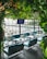 A modern outdoor café with vibrant green foliage hanging from the ceiling, offering a lush atmosphere. The area is furnished with sleek, blue-cushioned seating and glossy tables. Large glass windows provide a view of the waterfront and vehicles parked nearby.