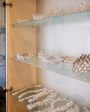 A glass display case is filled with an array of glittering tiaras and crowns, each intricately designed with crystals and pearls. The shelves showcase the delicate craftsmanship of the jewelry pieces, which are neatly arranged with some necklaces and matching accessories.