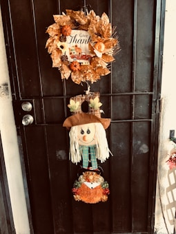 A decorative fall wreath adorned with orange pumpkins, golden leaves, and pinecones hangs on a black door. Below it, a cheerful scarecrow decoration features a smiling face with blonde straw hair, wearing a plaid blue scarf. Further down, an orange and green glittery pumpkin basket completes the autumn-themed arrangement.