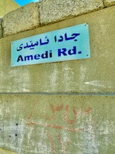 A blue metal sign attached to a concrete wall displays the text 'Amedi Rd.' in both English and Arabic script. The wall has a rough texture and appears weathered, with graffiti in red below the sign.